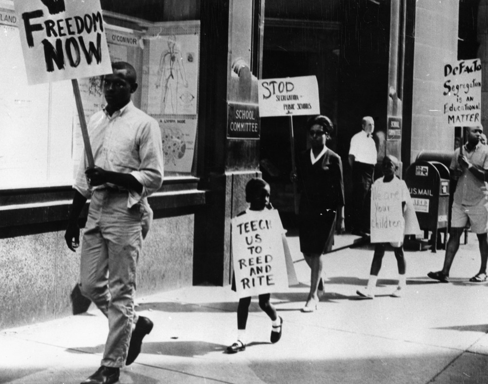 Black and white image of protestors holding pick signs while walking on a sidewalk