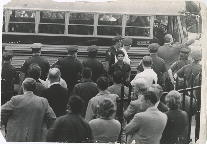 Black and white image of a student standing in front of a school bus surrounded by police and other people