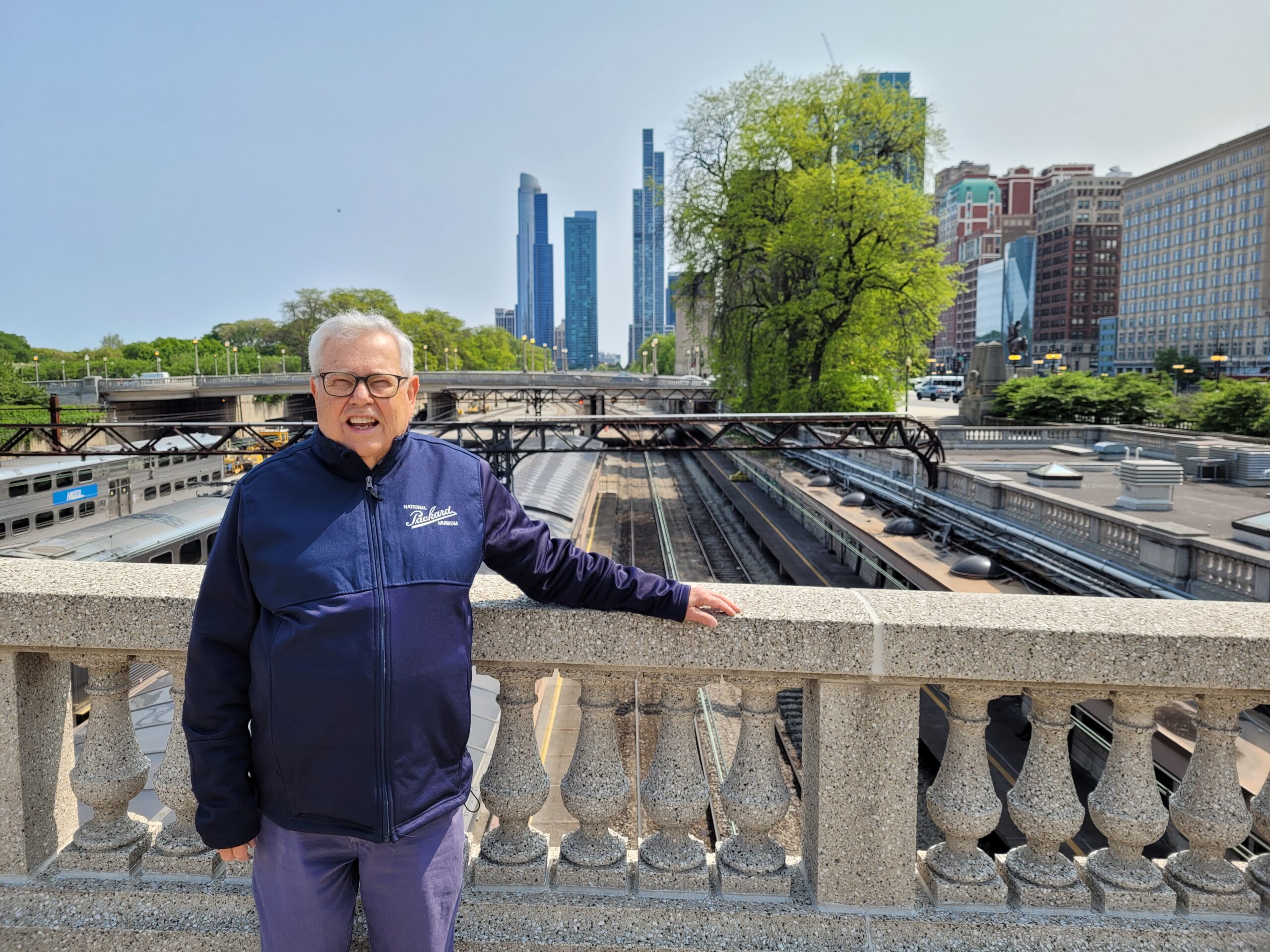 A smiling man wearing glasses and a blue sweatshirt stands on a bridge overlooking train tracks. Skyscrapers and trees are in the distance