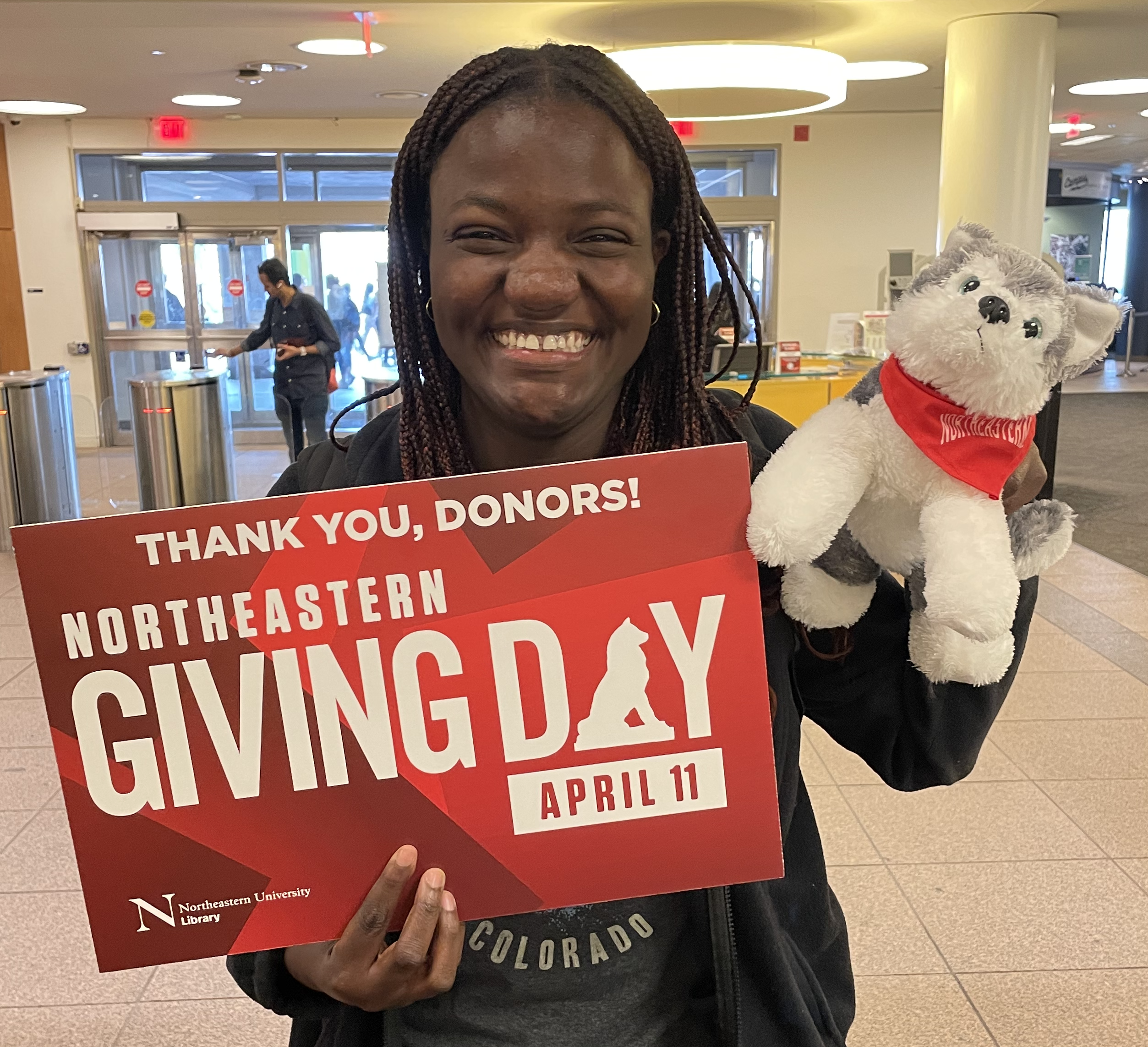 A student in Snell Library smiles while holding a "Thank you, donors!" Giving Day sign and a stuffed husky